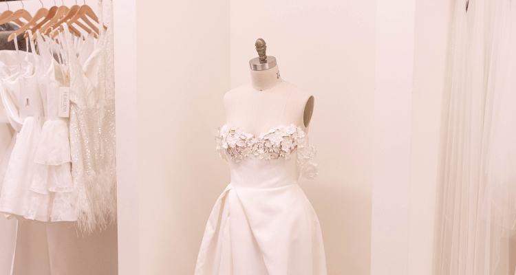 Elegantly tailored white dress with floral embellishments on a mannequin, surrounded by delicate, hanging white dresses in a bridal boutique.