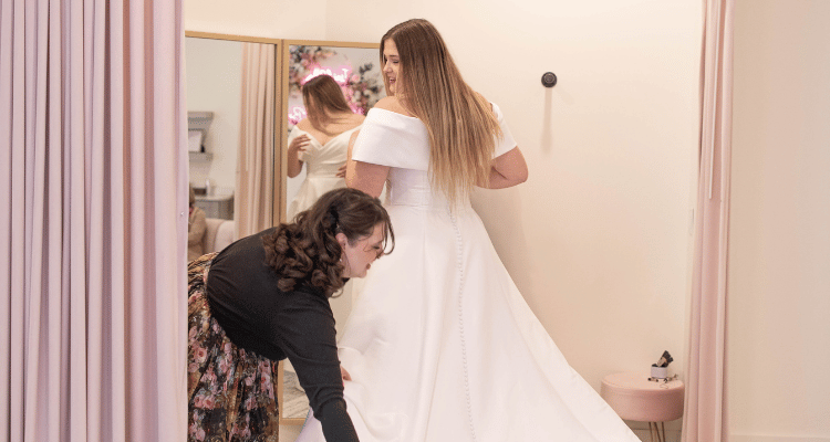 Bridal consultant adjusting the train of an off-the-shoulder wedding gown during a dress fitting appointment