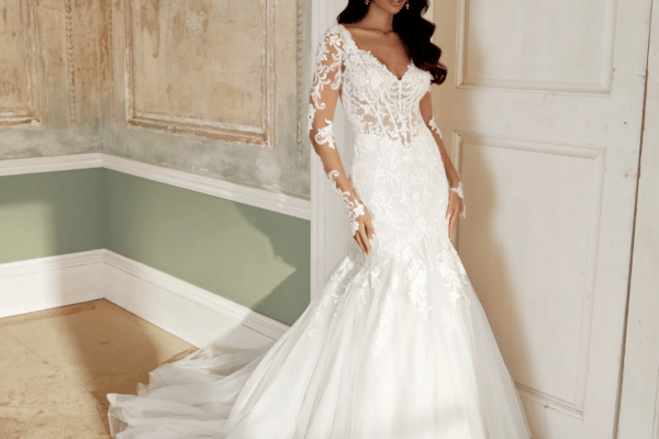 Bride in elegant wedding dress with lace sleeves stands in a vintage room. The gown's long train cascades beautifully, conveying romance and sophistication.