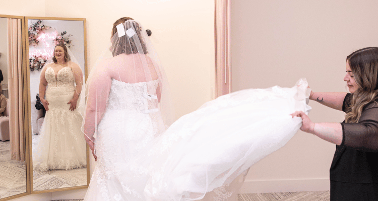 Bride viewing herself in a lace wedding dress while a bridal consultant arranges the train during a bridal appointment