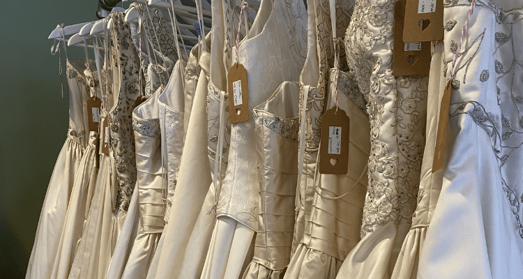 A row of elegant white and ivory wedding dresses with intricate lace and embroidery hang on a rack. Each garment has a tag attached, creating a luxurious and romantic atmosphere.