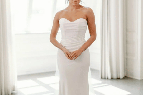 A woman stands in a sunlit room wearing a strapless white wedding dress, hands gently clasped. Soft curtains frame the serene, elegant scene.
