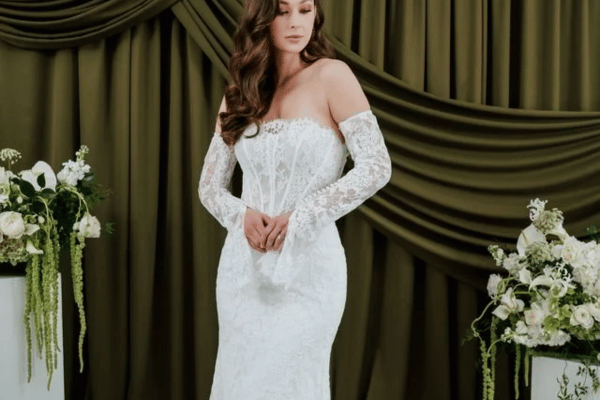 A woman in a lace bridal gown with sheer long sleeves stands elegantly against an olive-green backdrop. Flanking her are white floral arrangements.