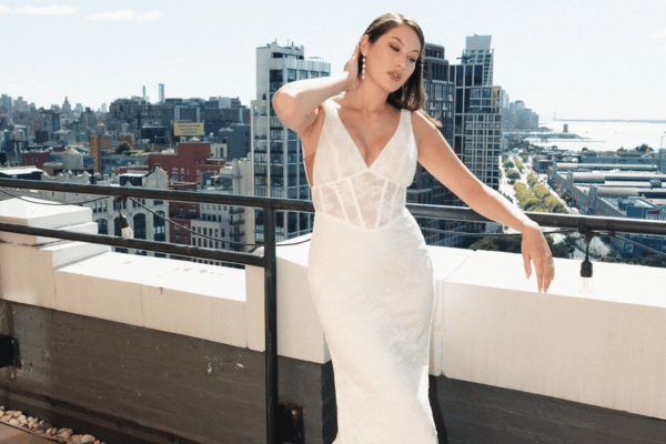 A woman in a flowing white dress poses elegantly on a rooftop, with a city skyline and blue sky in the background, exuding a serene and confident vibe.