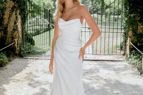 A woman in a white strapless gown stands in a sunlit path before an ornate iron gate. Lush greenery surrounds, exuding elegance and tranquility.