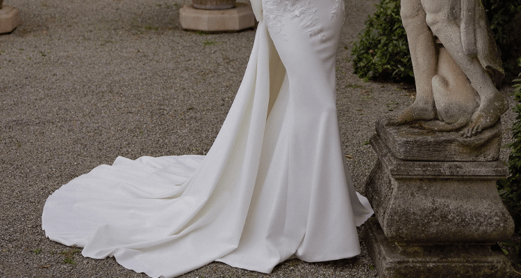 Bride wearing a fitted wedding dress with a long flowing train on a gravel path in a garden setting.