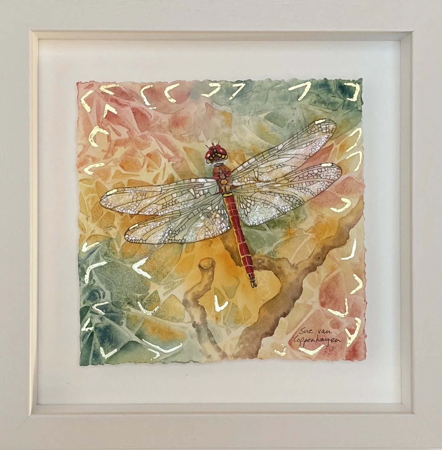 Painting of a red bodied dragonfly with shimmering wings against a semi abstract background in red, gold and green, with touches of gold leaf applied, in a square off white wooden frame