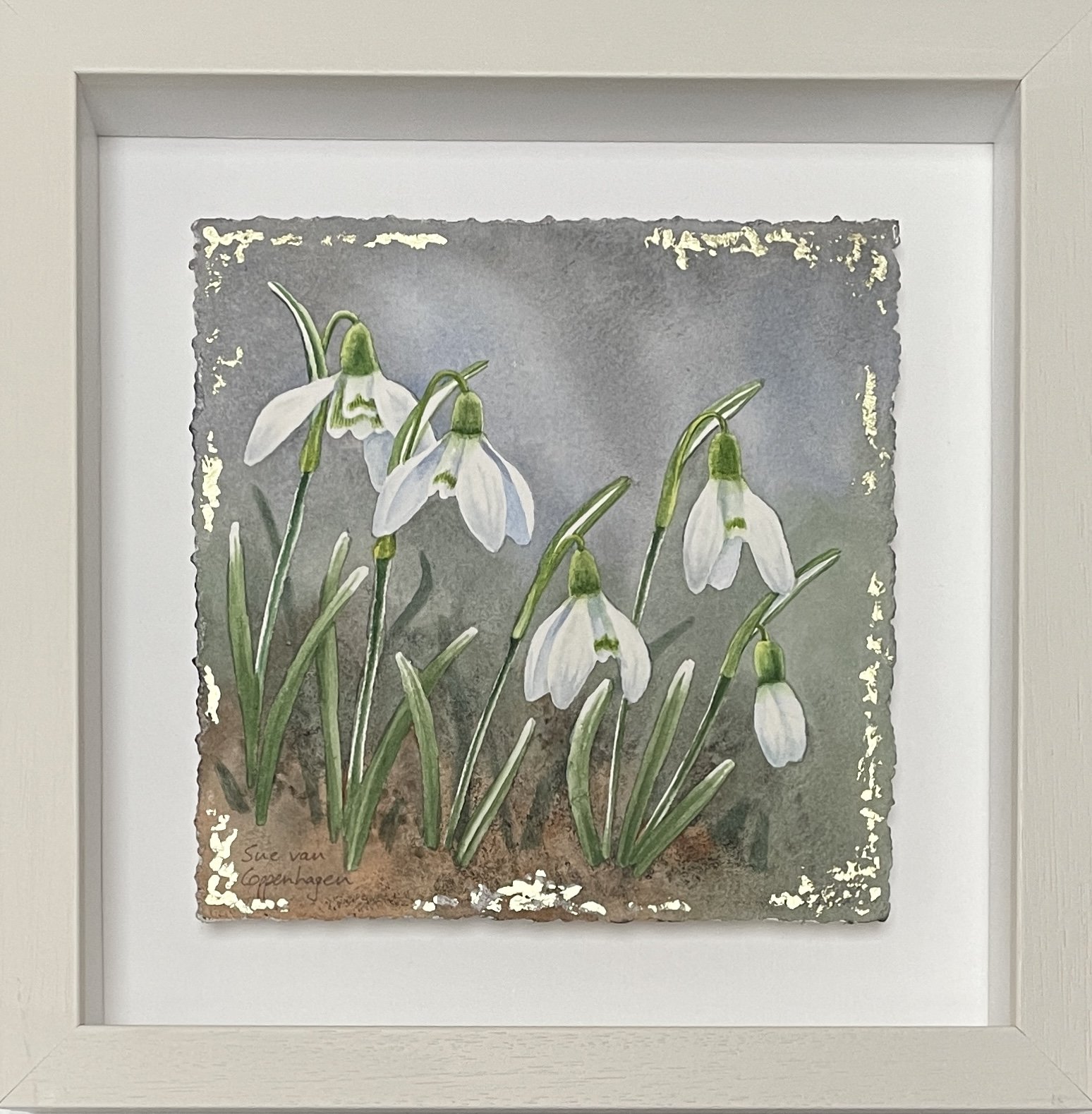 Painting of white snowdrop flowers with green leaves on a blue grey background edged with gold leaf, framed in a square off-white frame
