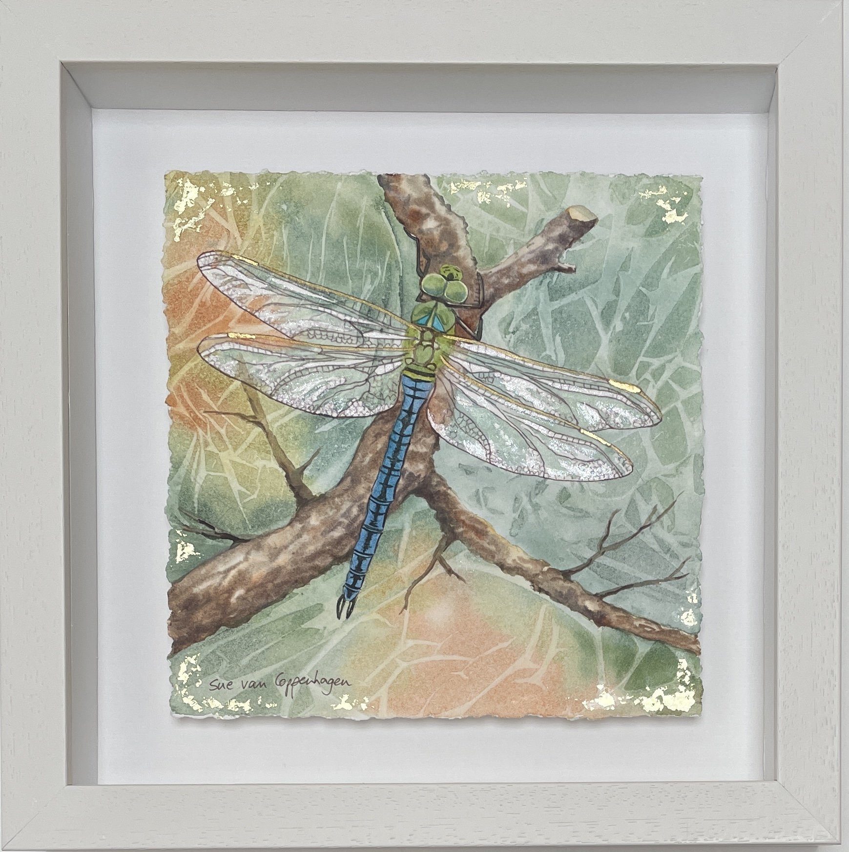 Painting of a shimmering dragonfly on a softly patterned green and copper background, framed in an off white wooden frame