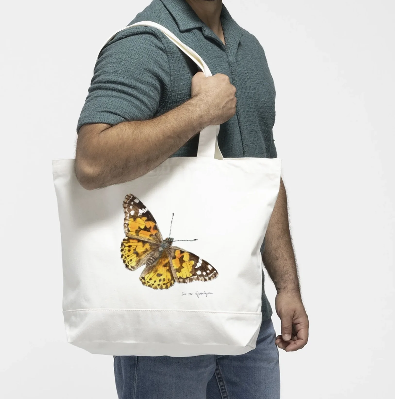 White cotton canvas tote bag with a painting of a Painted Lady butterfly ,modelled by a man in a blue shirt