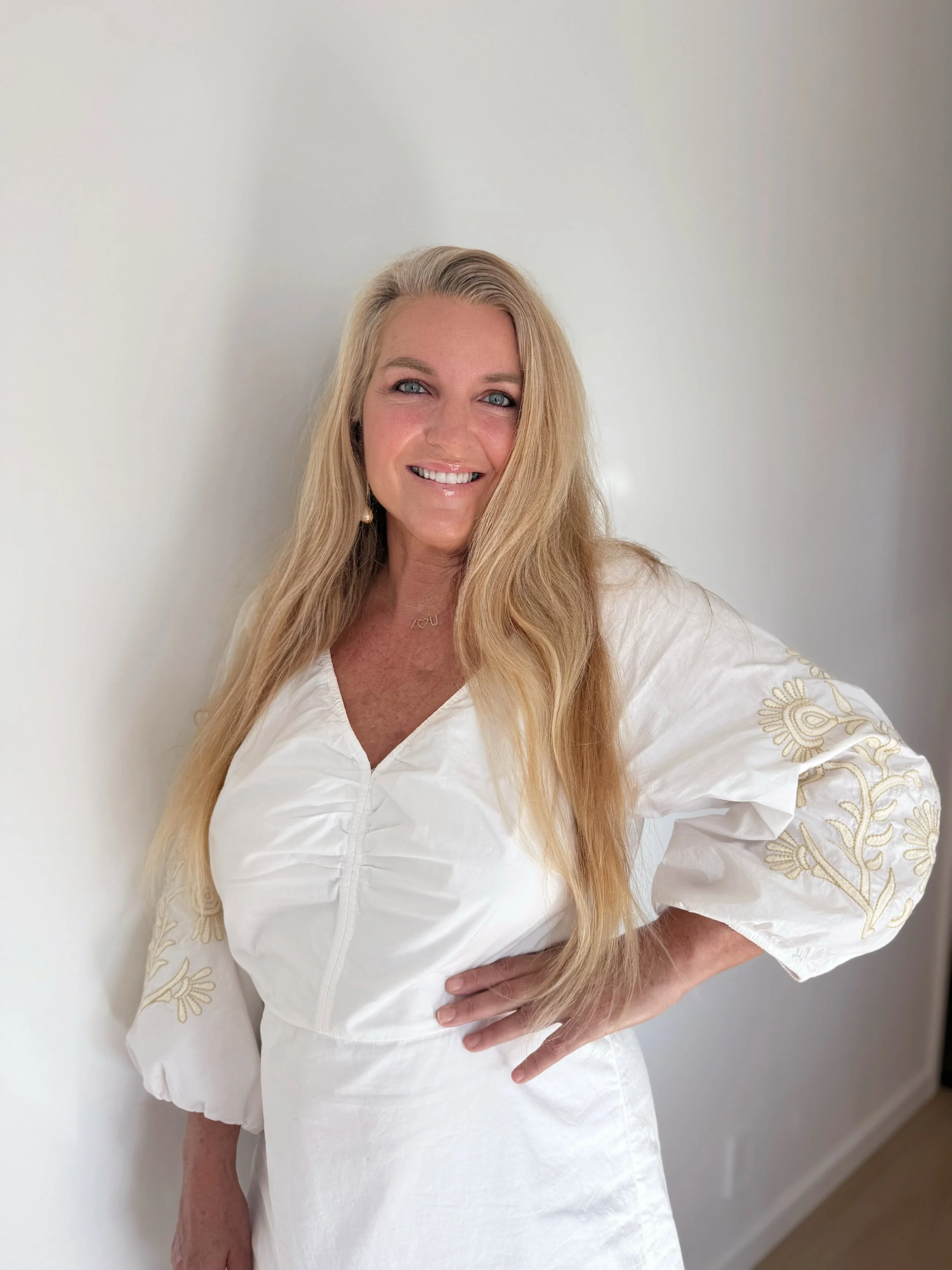 A woman with long blonde hair, smiling, wearing a white dress with gold embroidery on the sleeves, standing against a plain white wall.