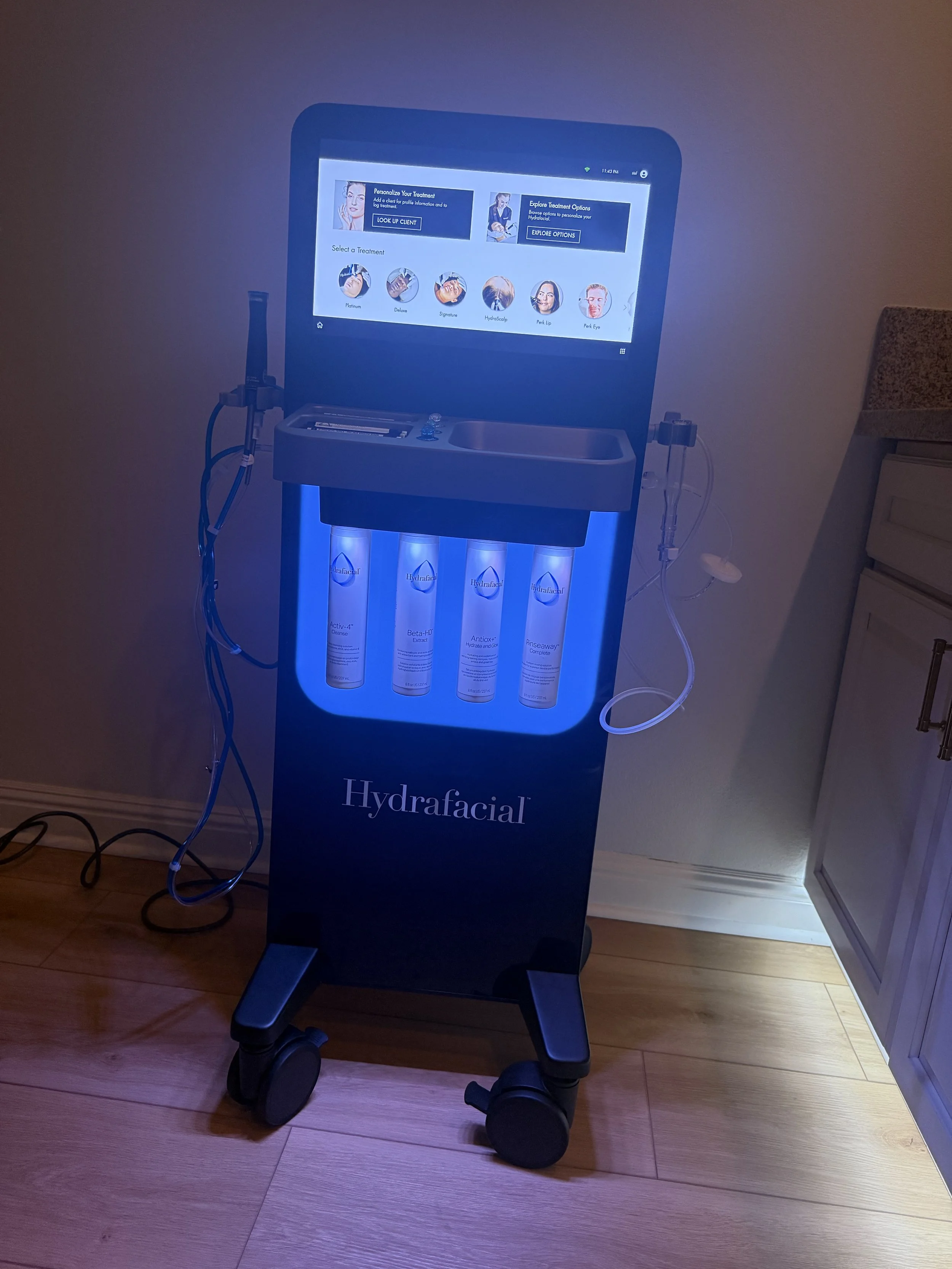 Hydrafacial machine with a touchscreen display showing treatment options and in-office skincare product bottles at the base.