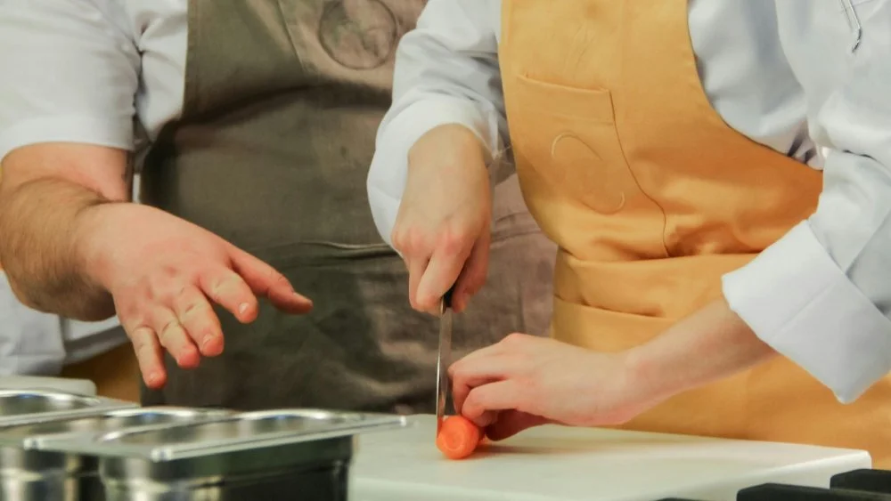 An instructor gives direction in how to properly chop a carrot while a student follows.