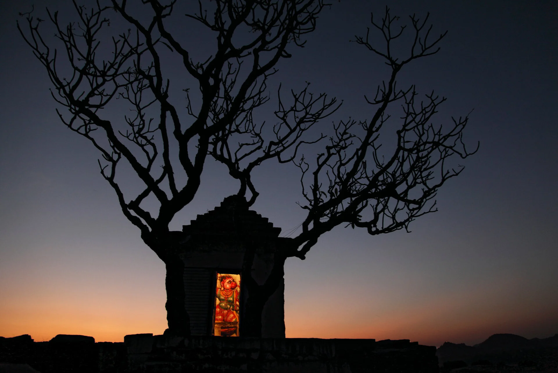 Silhouette eines Baumes und eines kleinen Altars mit einer goldenen hinduistischen Statue bei Sonnenuntergang im Hintergrund.
