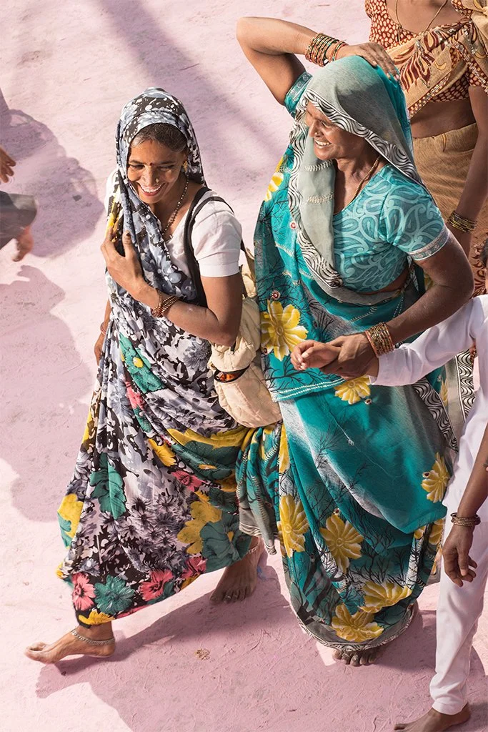 Zwei lachende Frauen in bunten Saris, die auf pinkem Sand tanzen und feiern.