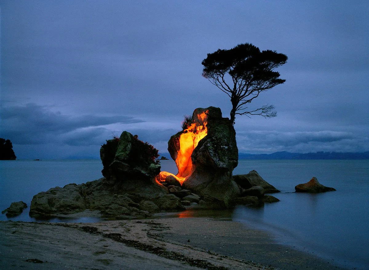 Ein Felsen im Wasser mit einem brennenden inneren Feuer, einem Baum auf dem Felsen, dunkler Himmel und Wasser im Hintergrund.