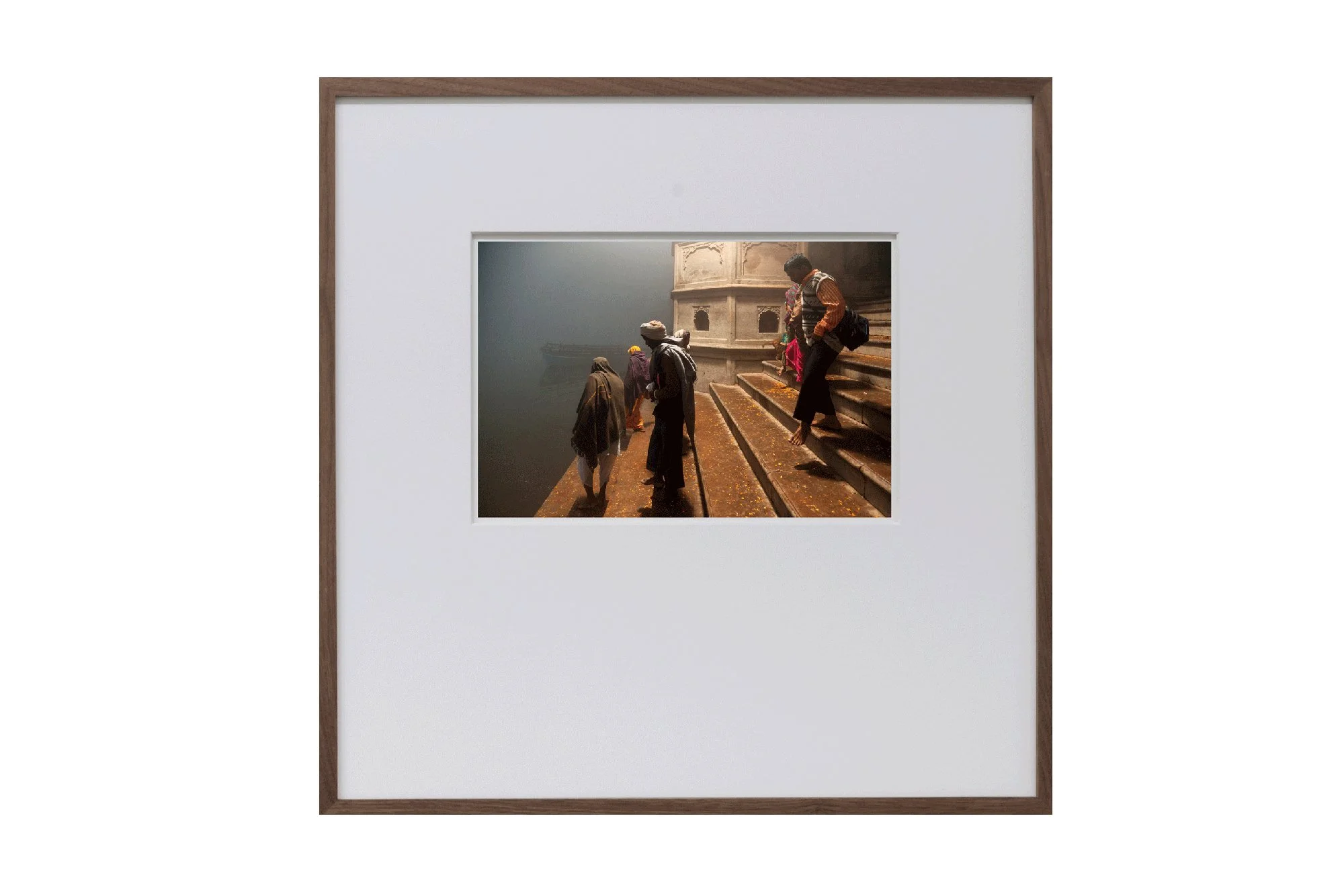 Fotografie von Menschen, die die Treppe am Ufer des Flusses Ganges in Indien hinuntersteigen, bei Nebel oder Rauch. Die Szene ist in einem Holzrahmen mit weißem Passepartout eingeschlossen.