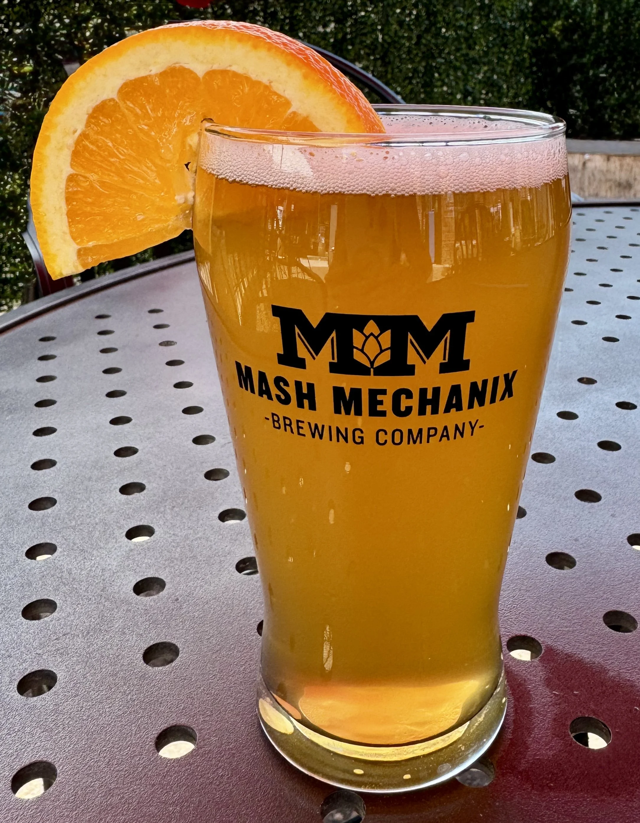 A pint of orange-hued wheat beer in a Mash Mechanix Brewing Company glass sits on a patio table, garnished with a fresh orange slice on the rim.