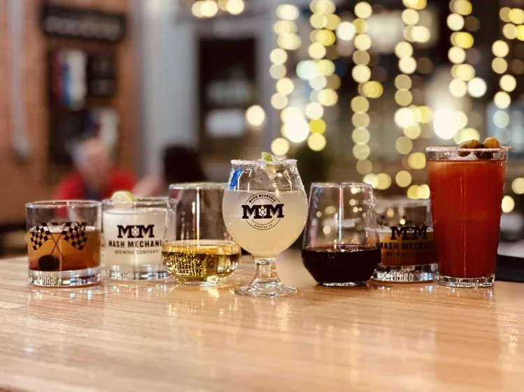 A lineup of cocktails and mixed drinks in Mash Mechanix Brewing glasses sits on a wooden bar with warm string lights glowing in the background.