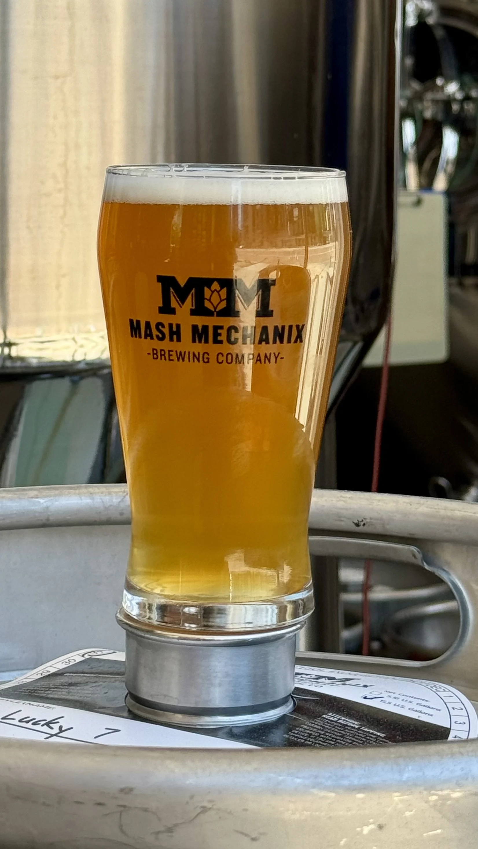 A pint of hazy golden Lucky 7 Hazy IPA in a Mash Mechanix Brewing Company glass sits on top of a brewery keg in front of stainless steel brewing tanks, with a light foamy head and the brewhouse equipment softly blurred in the background.