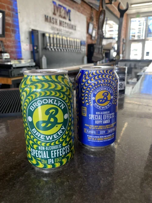 Two cans of non-alcoholic beer from Brooklyn Brewery—Special Effects IPA and Hoppy Amber—sit on a brewery bar with tap handles in the background.