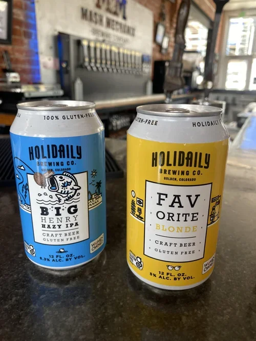 Two cans of gluten-free beer from Holidaily Brewing Co.—Big Henry Hazy IPA and Favorite Blonde—sit on a brewery bar in front of tap handles