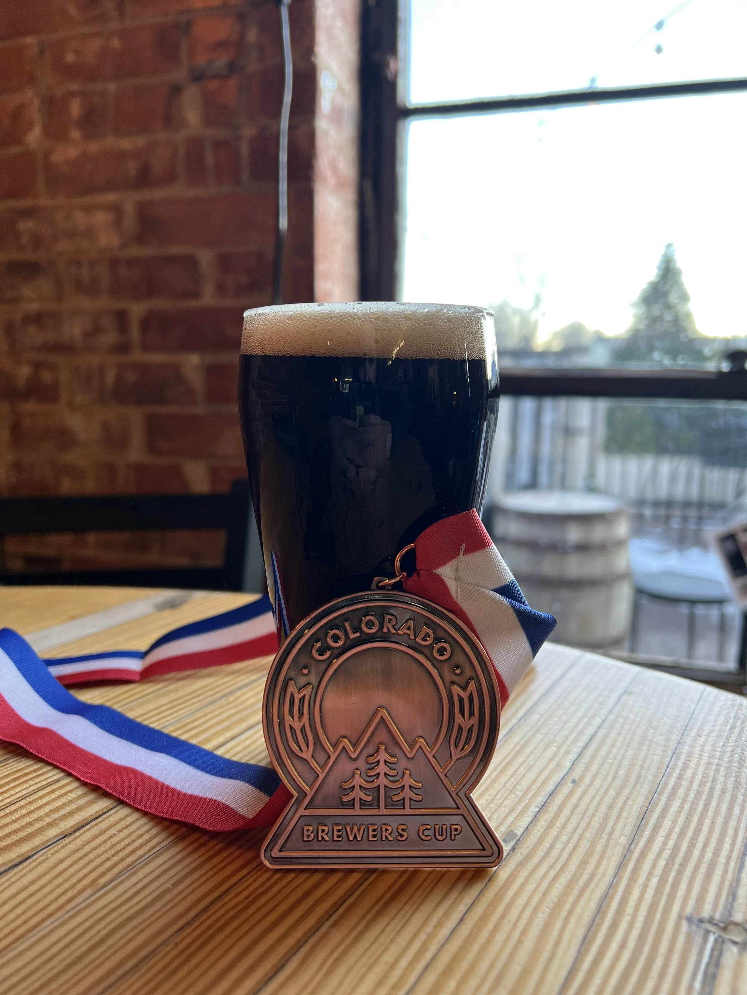 A pint of dark Power Brake Black IPA from Mash Mechanix Brewing sits on a wooden table next to a Colorado Brewers Cup medal with a red, white, and blue ribbon.