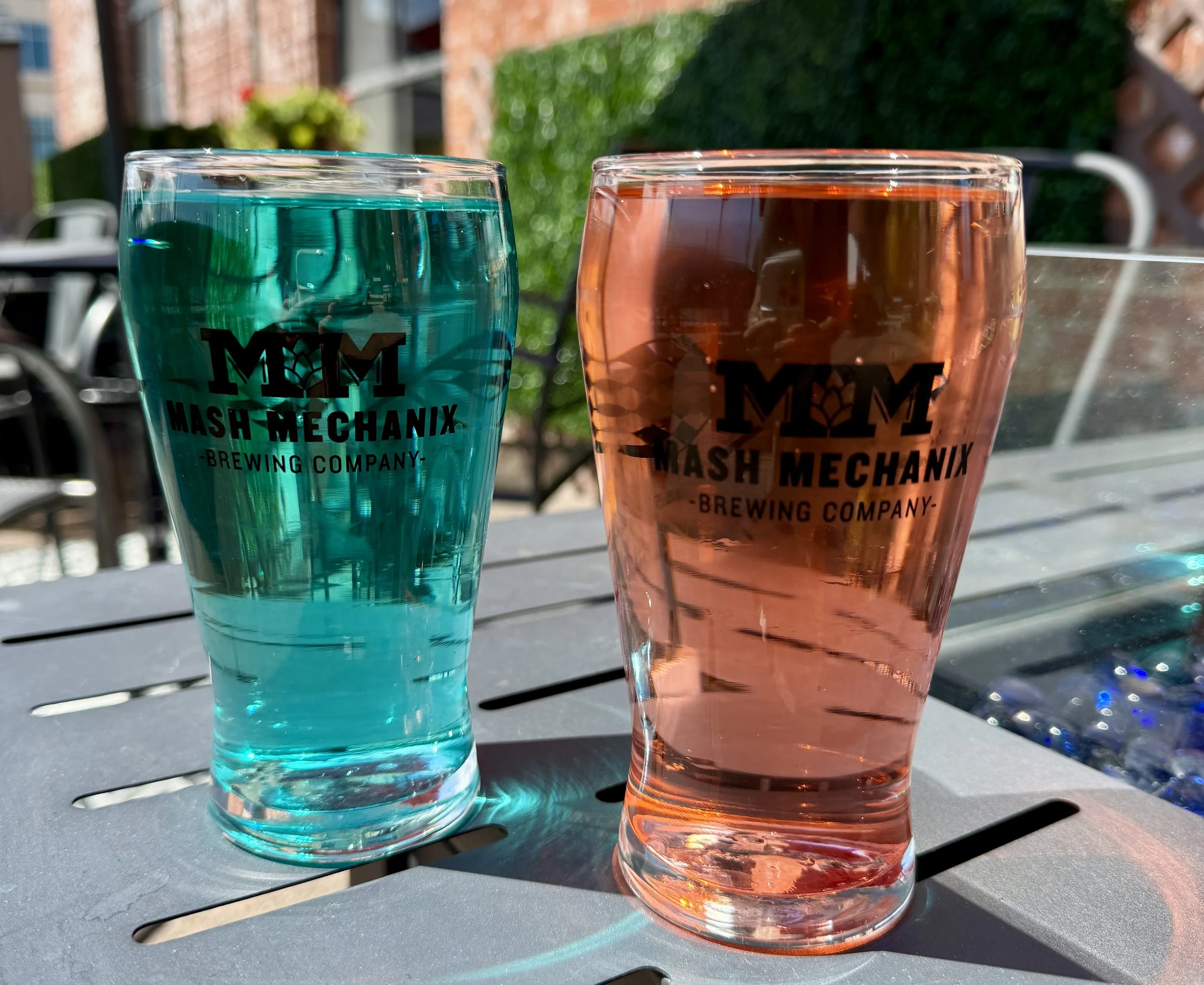 Two colorful hard seltzers in Mash Mechanix Brewing Company glasses—one bright blue and one pink—sit on an outdoor patio table in the sunshine.