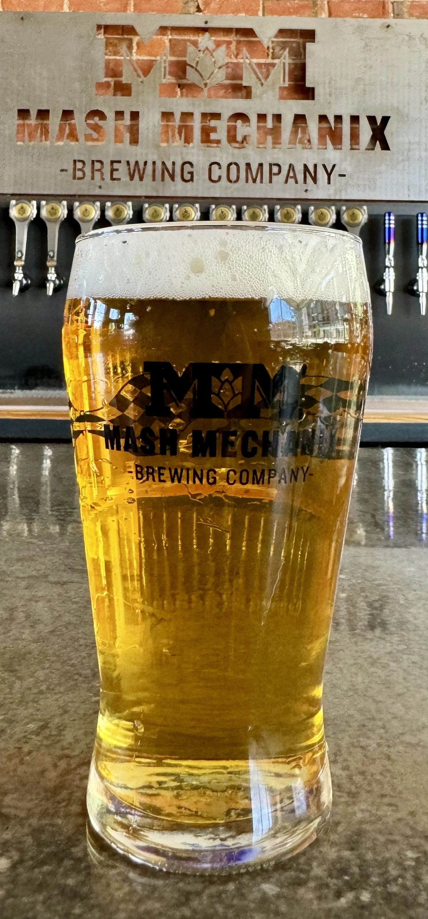 A pint of Larry’s Lager in a Mash Mechanix Brewing Company glass sits on the taproom bar, with a bright golden lager and a light foamy head, while the Mash Mechanix logo sign and a row of beer taps line the brick wall in the background.