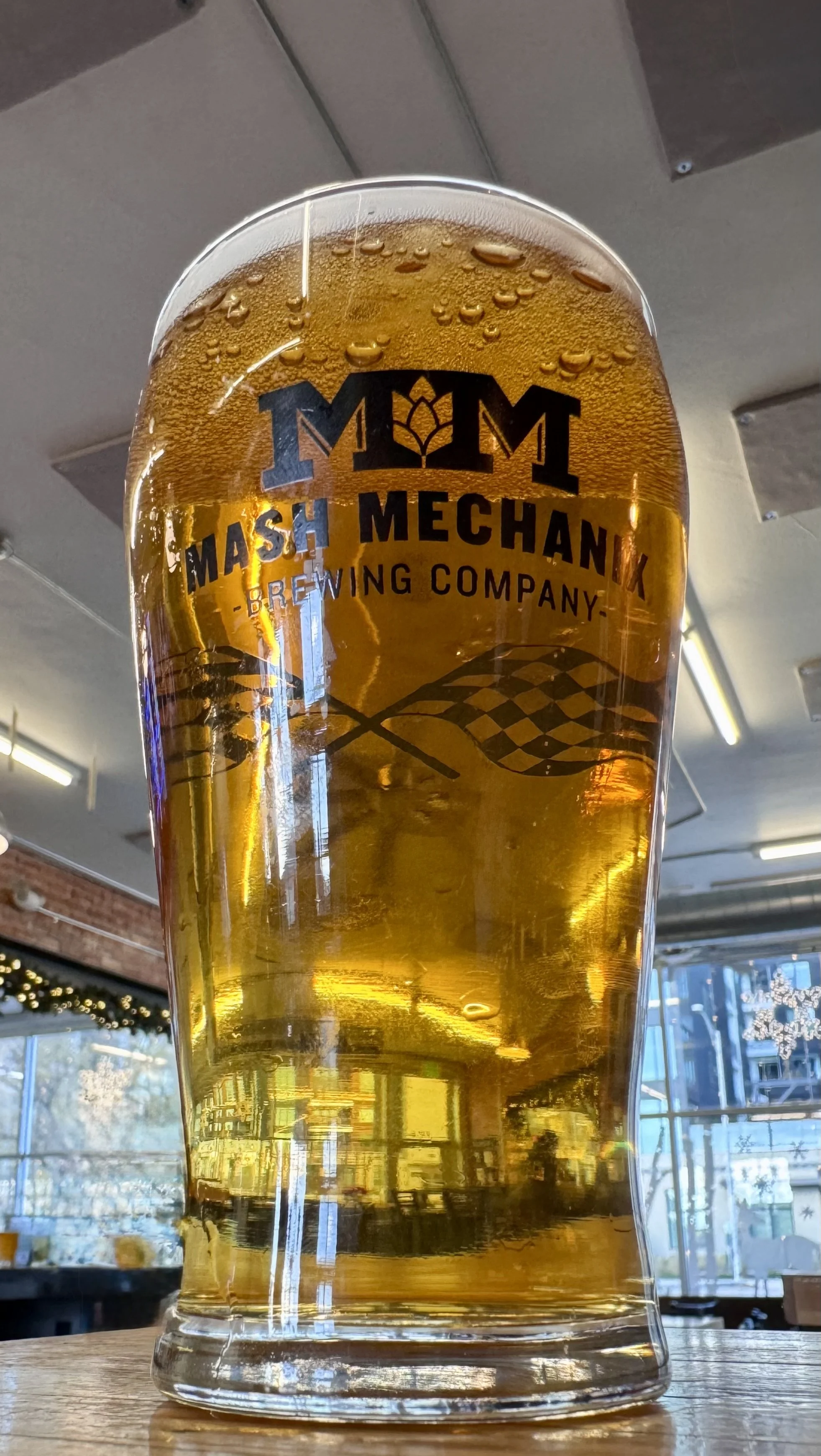 A pint of Sweet Ride Vanilla Cream Ale in a Mash Mechanix Brewing Company glass sits on a wooden table inside the bright taproom, showing a clear golden beer with rising bubbles and the brewery logo on the glass, with windows and warm lighting.