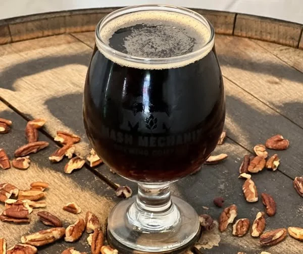 A glass of dark pecan brown ale from Mash Mechanix Brewing Company sits on a wooden barrel top surrounded by scattered pecans.