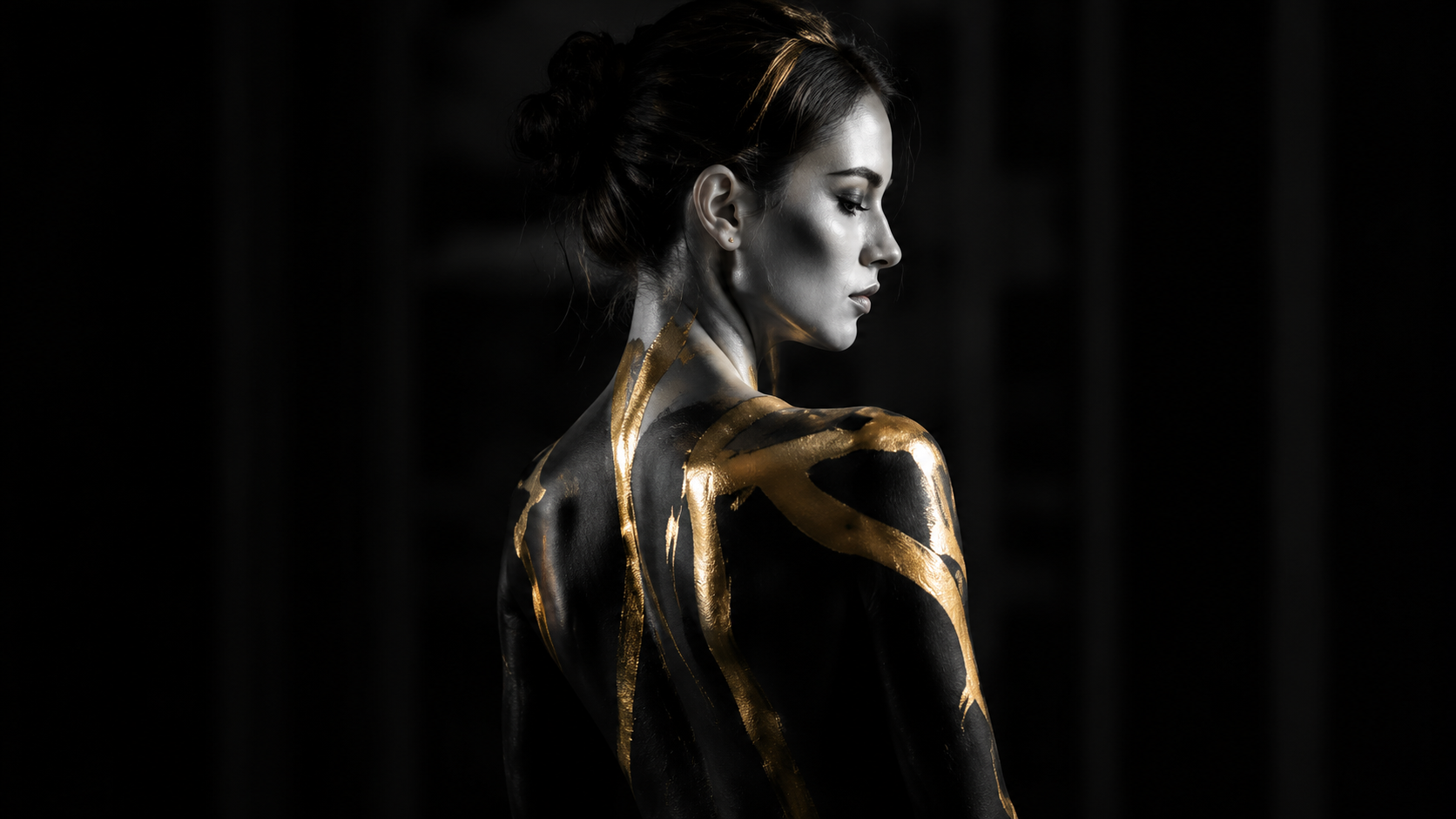 A woman with dark hair styled in an elegant updo, painted with gold streaks on her shoulder and back, posed against a dark background with soft lighting highlighting her profile and arm.