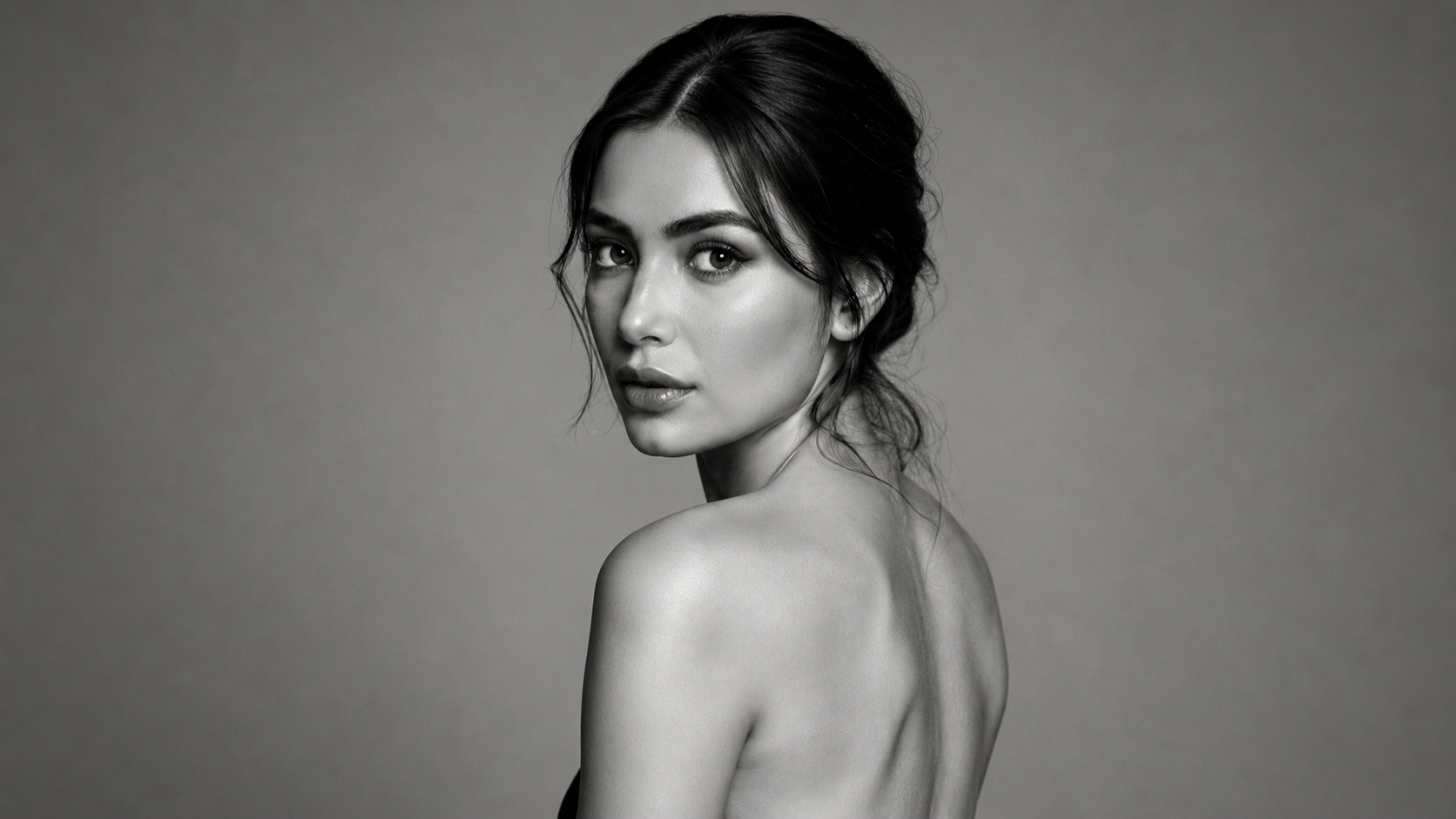 A black and white portrait of a young woman with dark hair, turned slightly to her right, looking into the camera, with bare shoulders.