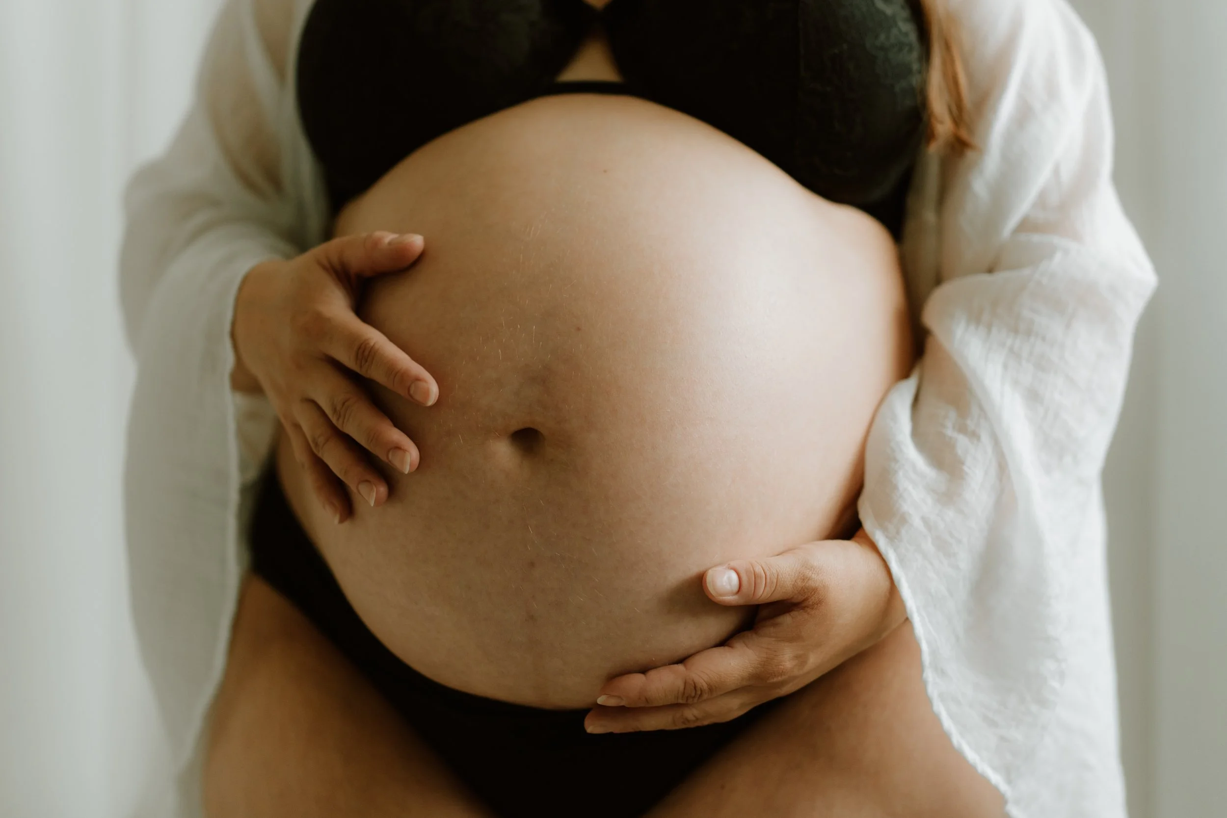 a pregnant woman in squamish, bc is posing for a maternity photoshoot, in a natural light studio. she has neatly filed nails, peach fuzz on her belly, and she's wearing a black bra with white linen robe