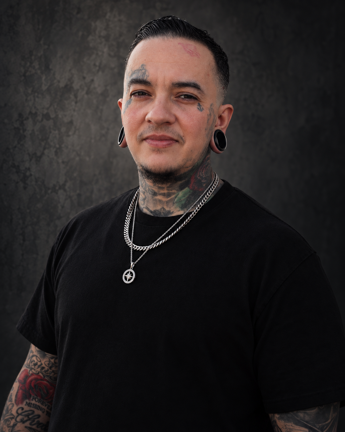 Portrait of a young man with dark hair and extensive tattoos, wearing a black t-shirt, layered silver necklaces, large black-rimmed earrings, looking at the camera against a dark textured background.