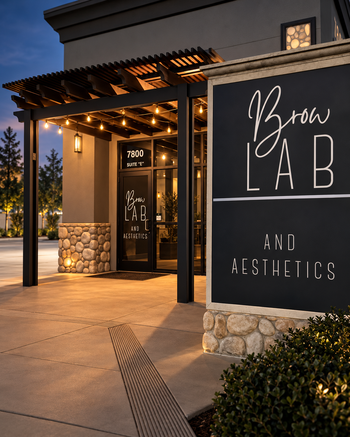 Exterior view of Brow's Lab and Aesthetics building at dusk with string lights hanging from the roof, a stone planter, and a black sign with white lettering.