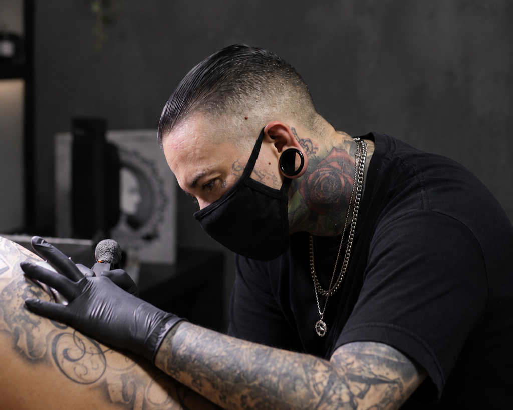 Tattoo artist wearing a black mask and gloves, working on a tattoo on a person's arm, in a tattoo studio.