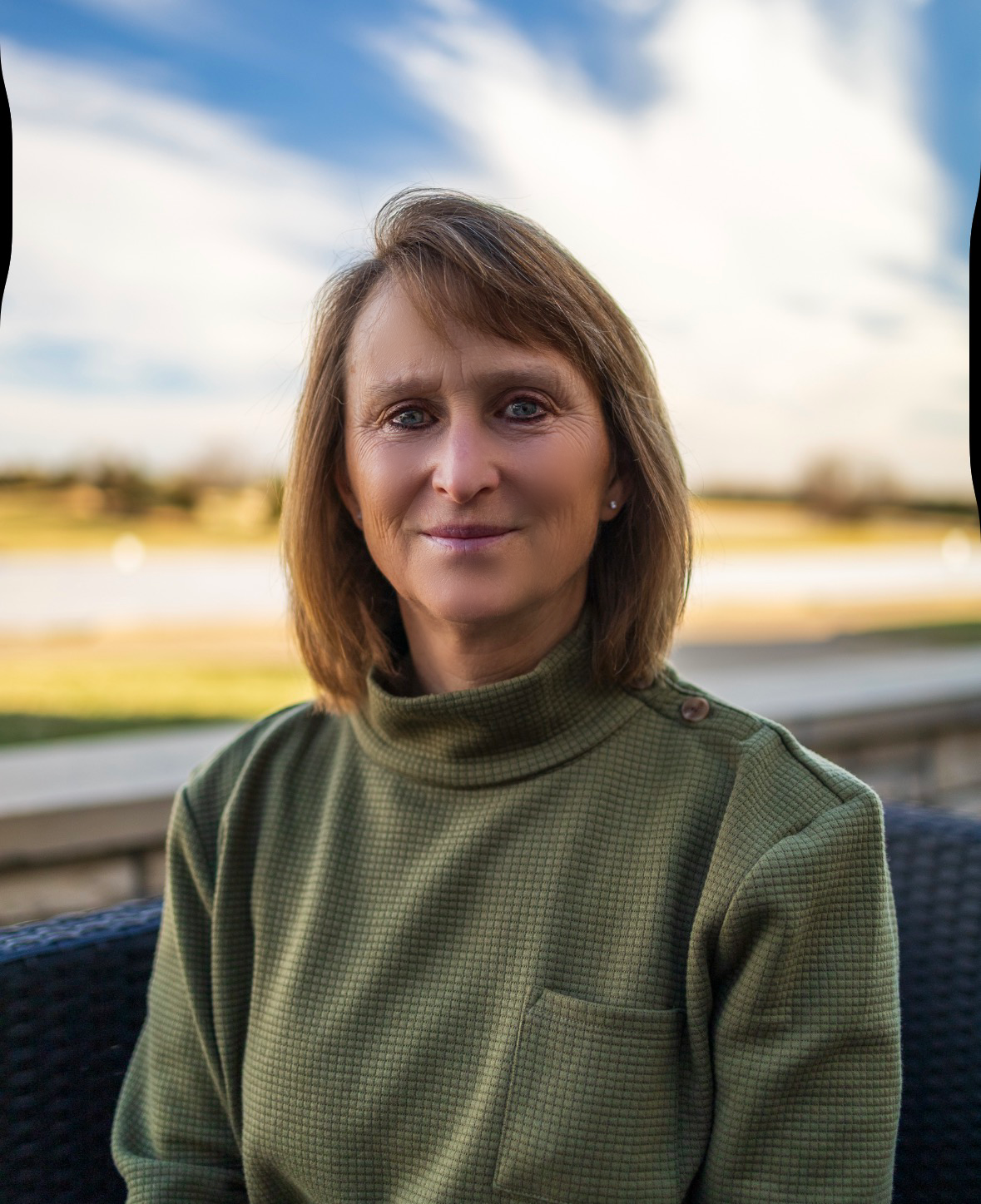 A woman with shoulder-length brown hair and blue eyes, wearing a green turtleneck sweater, sitting outdoors on a patio with a park and cloudy sky in the background.