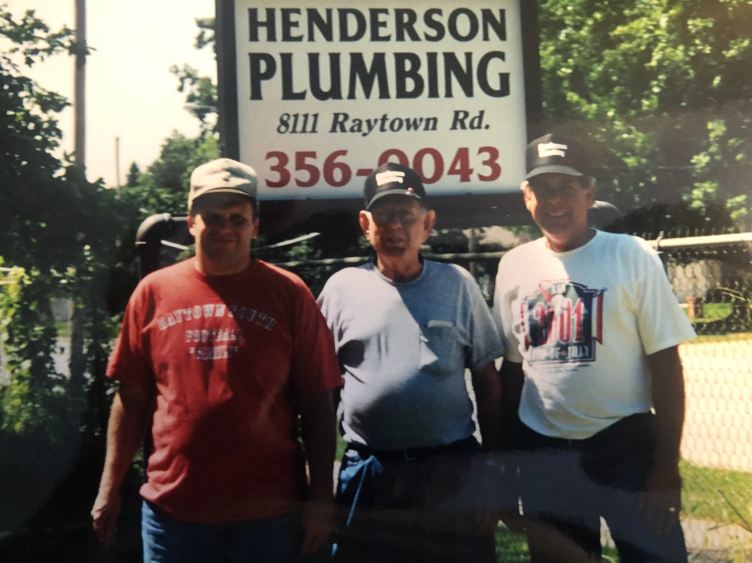 Three men standing outdoors in front of a Henderson Plumbing sign, wearing casual clothes and hats, smiling at the camera.