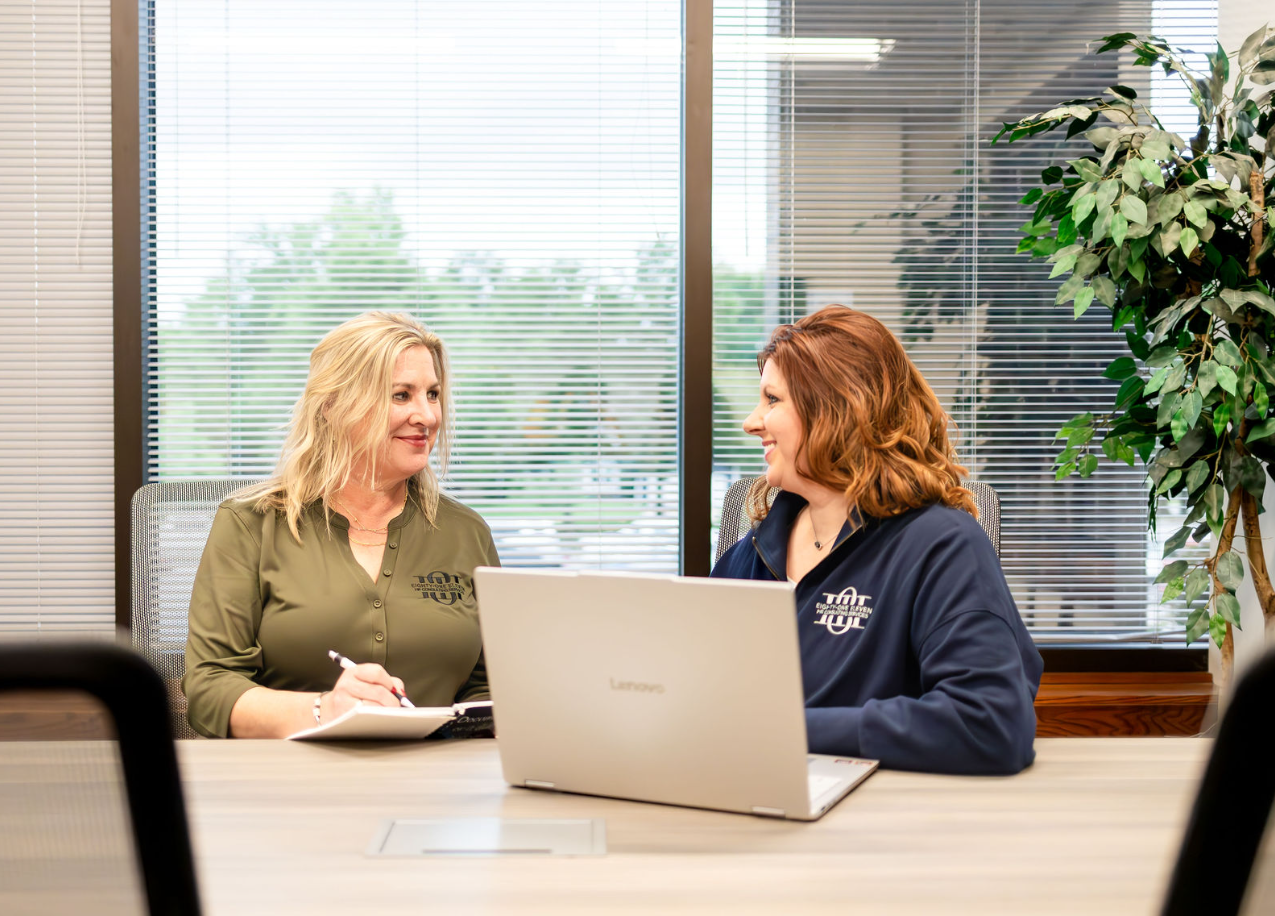 Two women having a discussion in a conference room, one with blonde hair and the other with red hair, sitting at a table with a laptop and notebook.