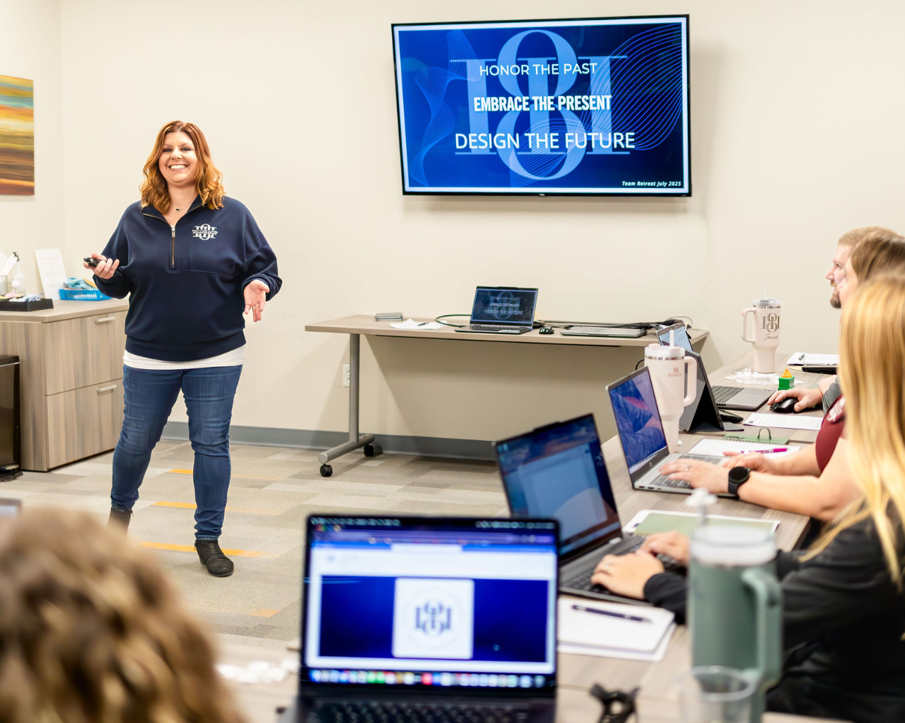 A woman giving a presentation in a conference room with a large screen displaying motivational phrases. She is smiling and holding a remote, and there are several attendees working on laptops at the table.