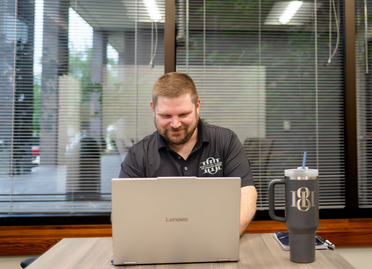 A man working on a Lenovo laptop at a desk in an office with large windows and blinds, with a travel mug and a smartphone on the desk.