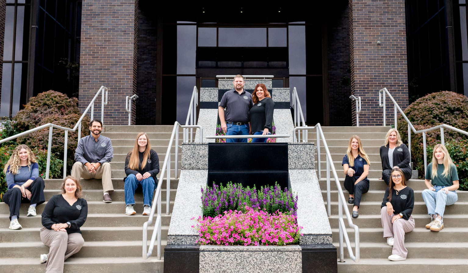 A group of ten people, including two standing on the top step, sitting on the steps, and standing on the side, pose outside a modern building with brick and glass architecture, and a flower bed with pink and purple flowers in the foreground.