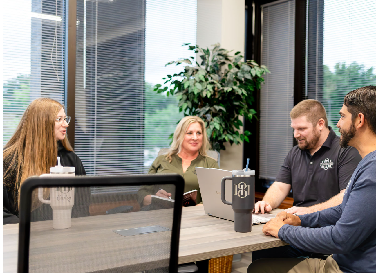 Four people sitting around a conference table engaged in a discussion in a modern office with large windows, blinds, and a large plant in the background.