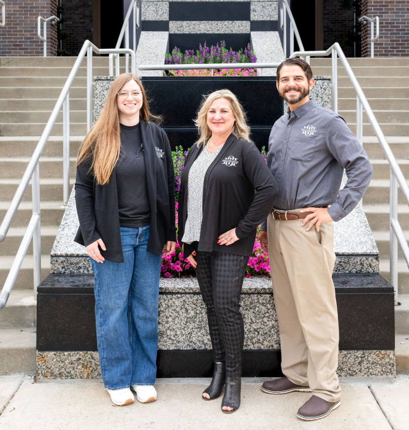 Three people standing in front of a staircase and flower bed, smiling at the camera.