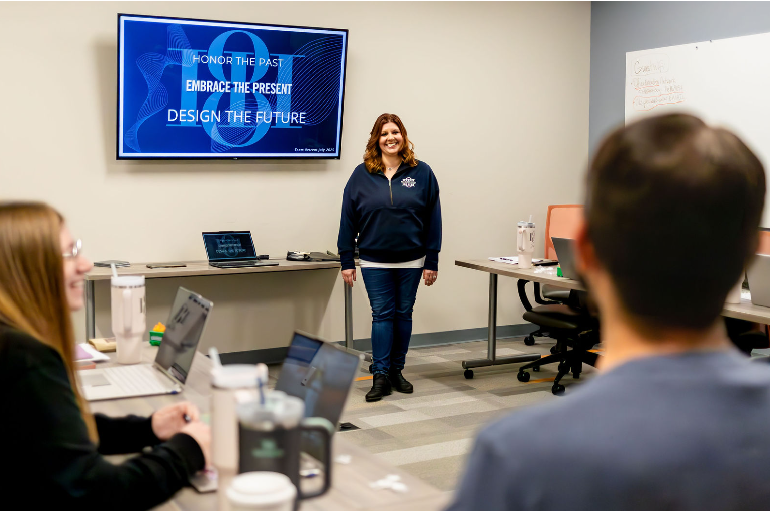 A woman stands in front of a presentation screen with the words 'Honor the Past, Embrace the Present, Design the Future' in a meeting room. She is smiling, wearing a navy jacket, and facing the seated group of people with laptops.