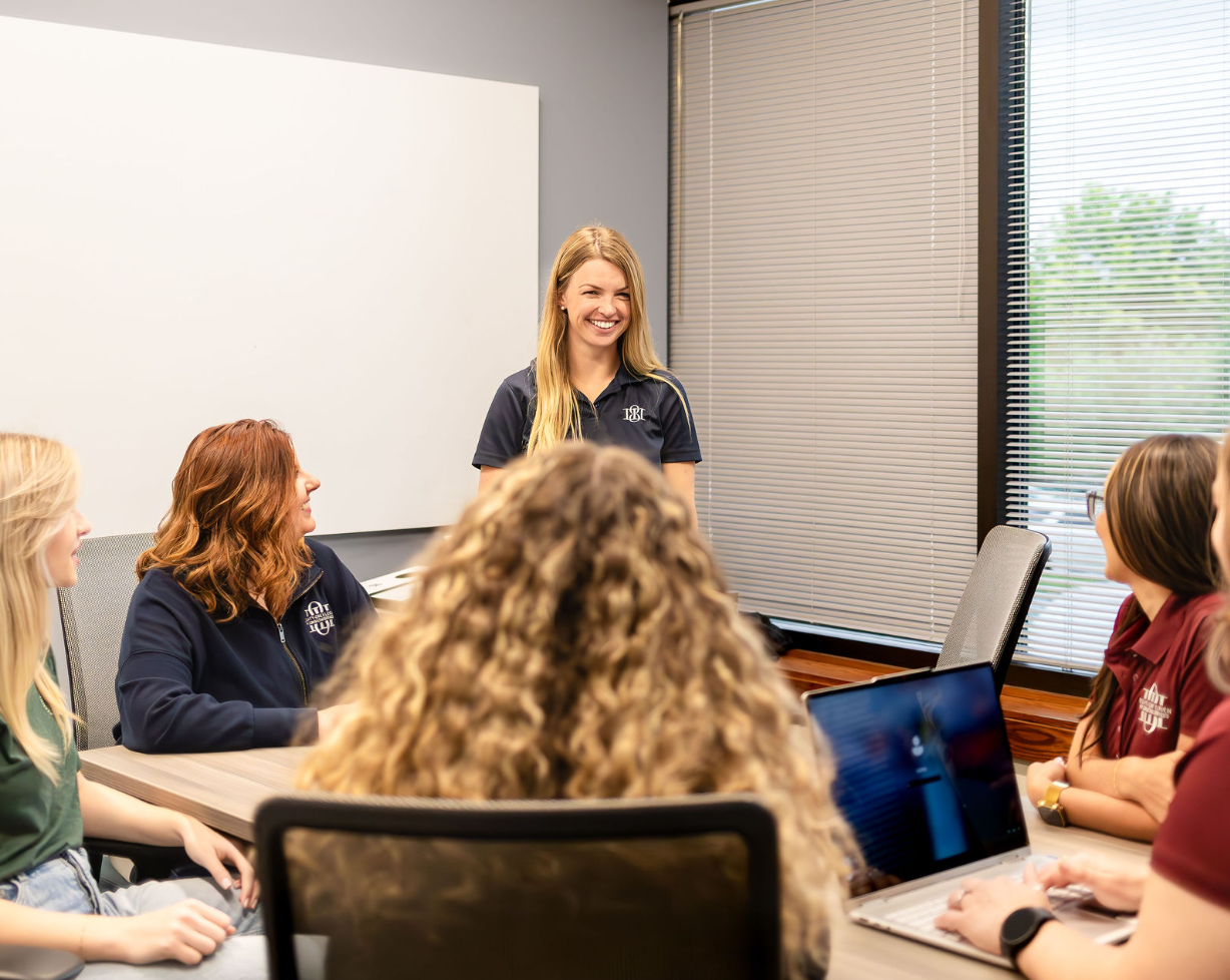 A group of women sitting around a conference table, one woman standing and smiling during a meeting in a bright office with large windows and blinds.