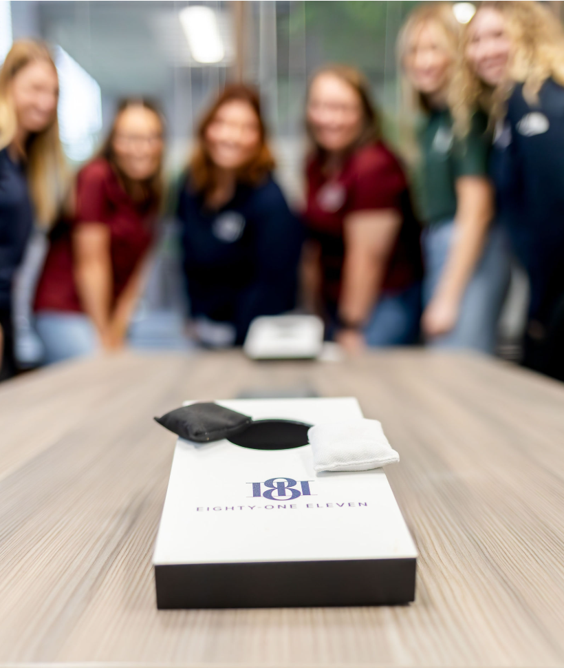 A box on a table with a black eye mask and a white cloth in the foreground, and a group of six people in the background, out of focus, smiling.