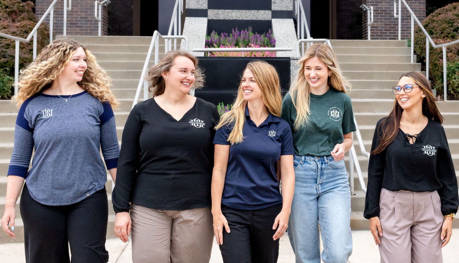 Five women walking together outside, smiling, in front of a building with stairs and flower boxes, wearing casual clothing with school or team logos.