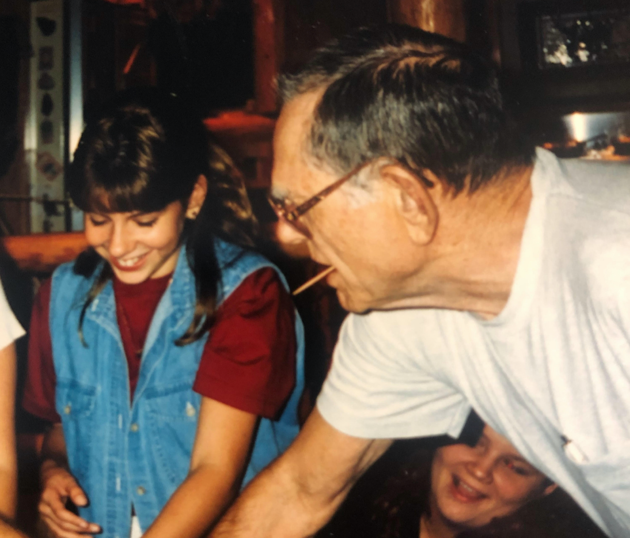 A man wearing glasses and a woman with brown hair and bangs smiling at a table, engaging in a shared activity, with another woman smiling in the background.